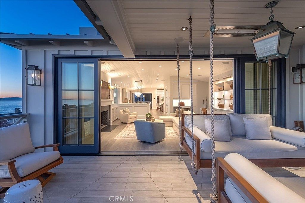 This image captures a seamless transition between a covered outdoor patio and a bright, open-concept living area. The patio features a hanging daybed, a lounge chair, and wood-look tile flooring, while the interior space showcases a fireplace, comfortable seating, and a view of the ocean through large glass doors. The design emphasizes indoor-outdoor living, with a neutral color palette and sophisticated coastal aesthetic.