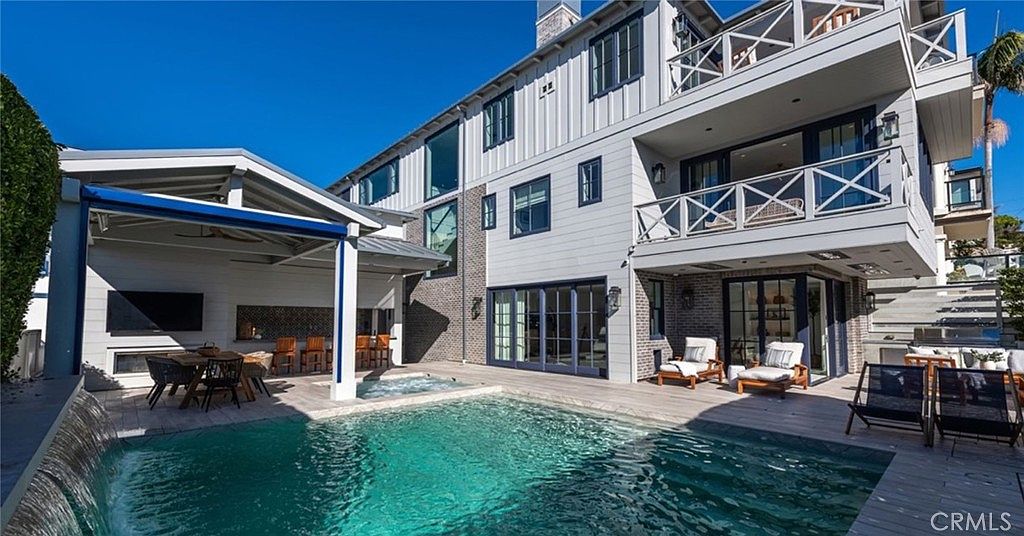 This luxurious backyard features a custom swimming pool with a built-in spa and a cascading water feature. A covered outdoor kitchen area with bar seating and a television provides an ideal space for entertaining, while a second-story balcony overlooks the patio. The home showcases a modern farmhouse aesthetic with light-colored siding, dark window frames, and brick accents, creating a sophisticated and inviting outdoor living environment.