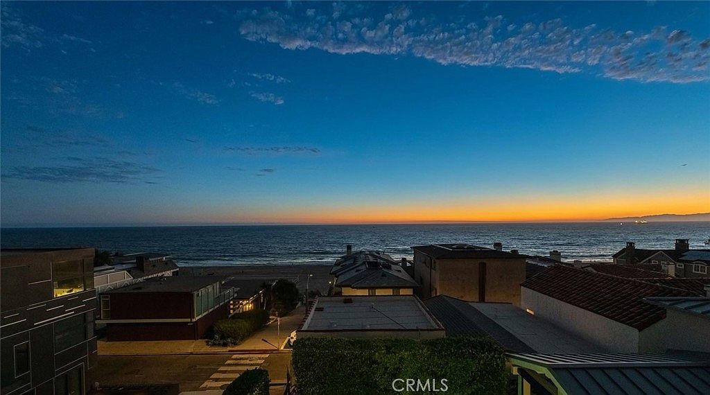 This elevated perspective captures a stunning sunset view over the ocean from a residential neighborhood. The scene features a coastal street lined with homes, leading toward a sandy beach and the vast horizon. The composition emphasizes the serene atmosphere and the prime location of the property, highlighting the transition from the quiet residential area to the expansive shoreline.