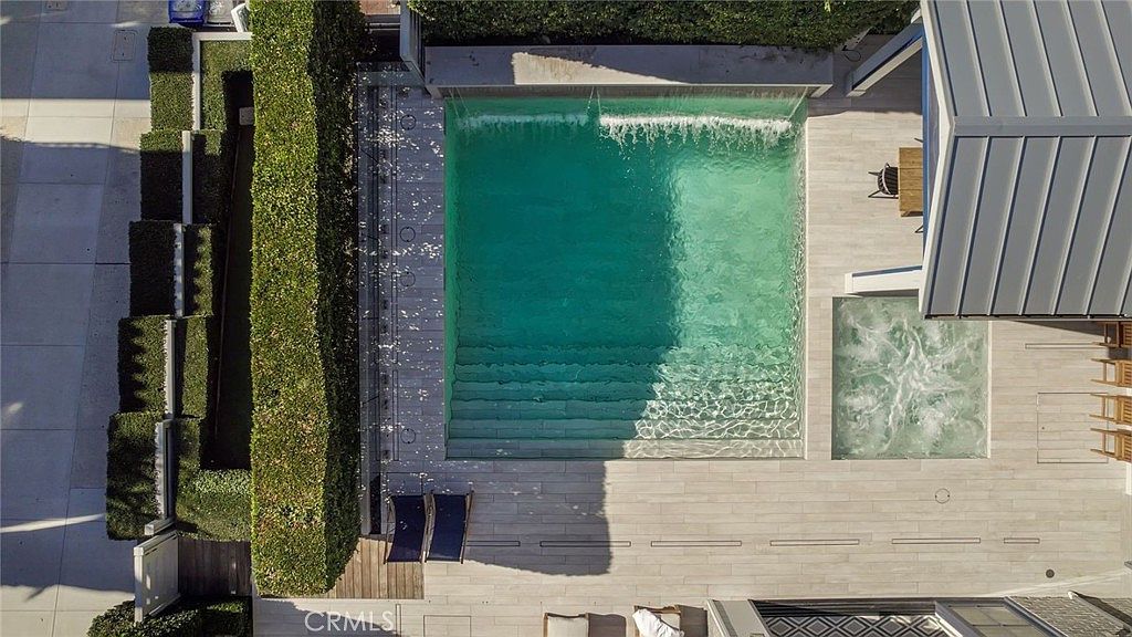 This high-angle aerial view captures a modern, rectangular swimming pool integrated with a square spa, both featuring light-toned stone decking. A cascading water feature flows into the pool from the rear wall, while a tall, manicured hedge provides privacy along the side. The clean lines and neutral color palette create a sophisticated, resort-like atmosphere perfect for outdoor relaxation.