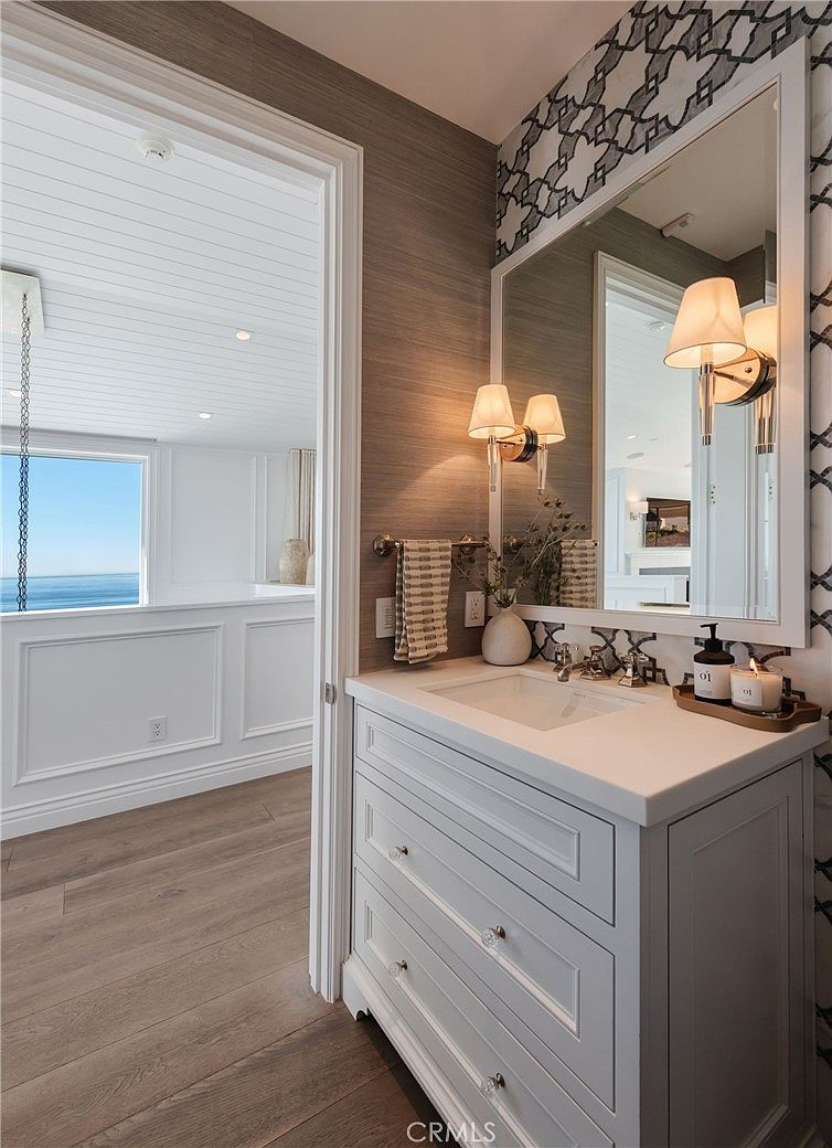 This elegant powder room features a white vanity with a stone countertop and a large, framed mirror set against a textured wall covering. The space is accented by dual wall-mounted light fixtures and a patterned wallpaper border above the mirror, creating a sophisticated aesthetic. A doorway provides a view into an adjacent room with light wood flooring and a white wainscoted wall overlooking the ocean.