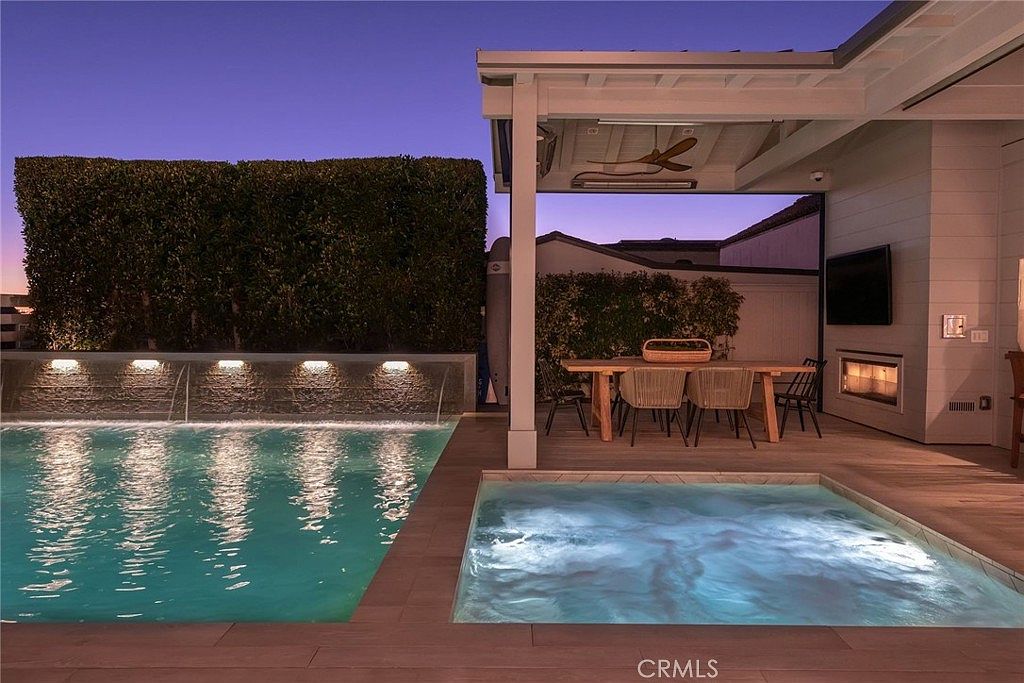 This evening shot captures a luxurious outdoor living space featuring a rectangular swimming pool with a water feature wall and an adjacent square spa. A covered patio area provides a comfortable dining space with a table and chairs, complemented by a built-in fireplace integrated into the wall. The scene is illuminated by soft ambient lighting, creating a serene and inviting atmosphere perfect for evening relaxation.