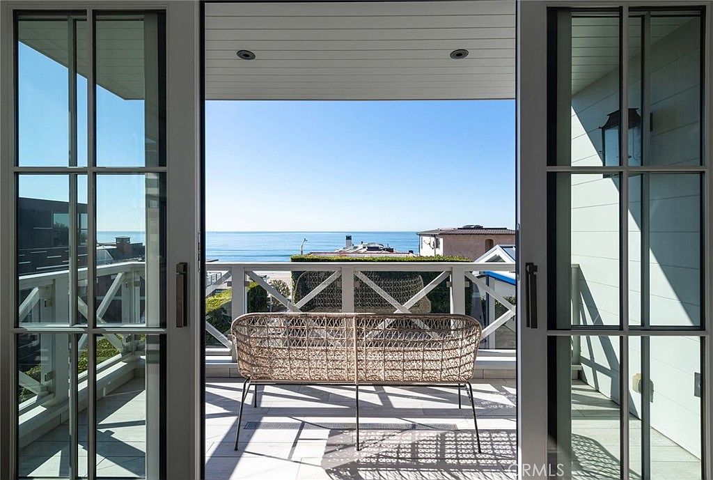 This image captures a serene balcony view looking out toward the ocean through large glass doors. The balcony features a woven bench seat placed on a patterned rug, creating a comfortable outdoor seating area. The perspective is from inside looking out, emphasizing the seamless transition between the indoor space and the expansive coastal horizon.