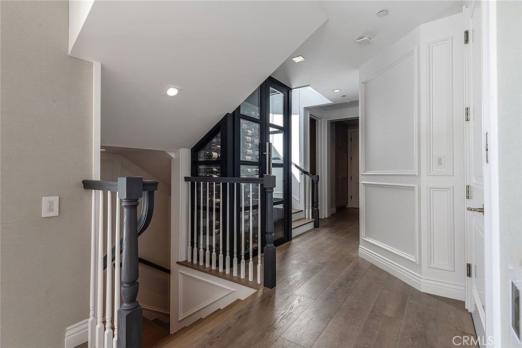 This interior view captures a sophisticated hallway featuring a staircase with dark-toned railings and white balusters. A prominent glass-enclosed wine storage unit with a black frame serves as a focal point, positioned near the transition to an upper landing. The space is finished with light wood flooring and white paneled walls, creating a bright and elegant atmosphere.