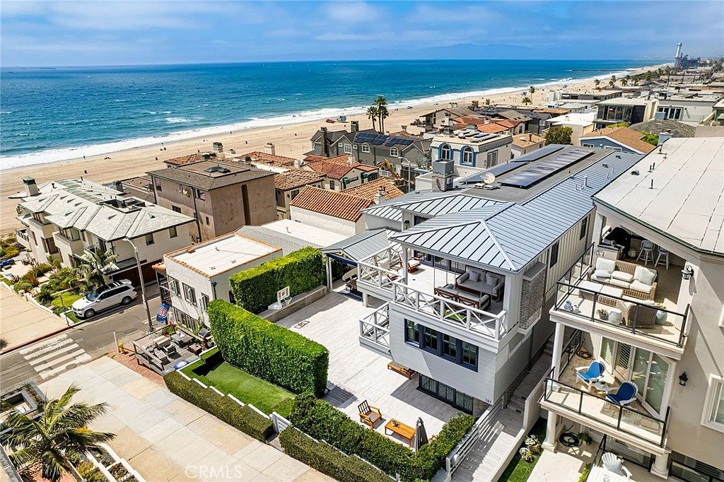This high-angle aerial view captures a coastal property situated just steps from the sandy beach and ocean. The home features a multi-level design with expansive decks, a private courtyard enclosed by tall hedges, and a modern aesthetic with light-colored siding and metal roofing. The perspective highlights the prime location, showcasing the seamless transition between the private outdoor living spaces and the surrounding beach community.