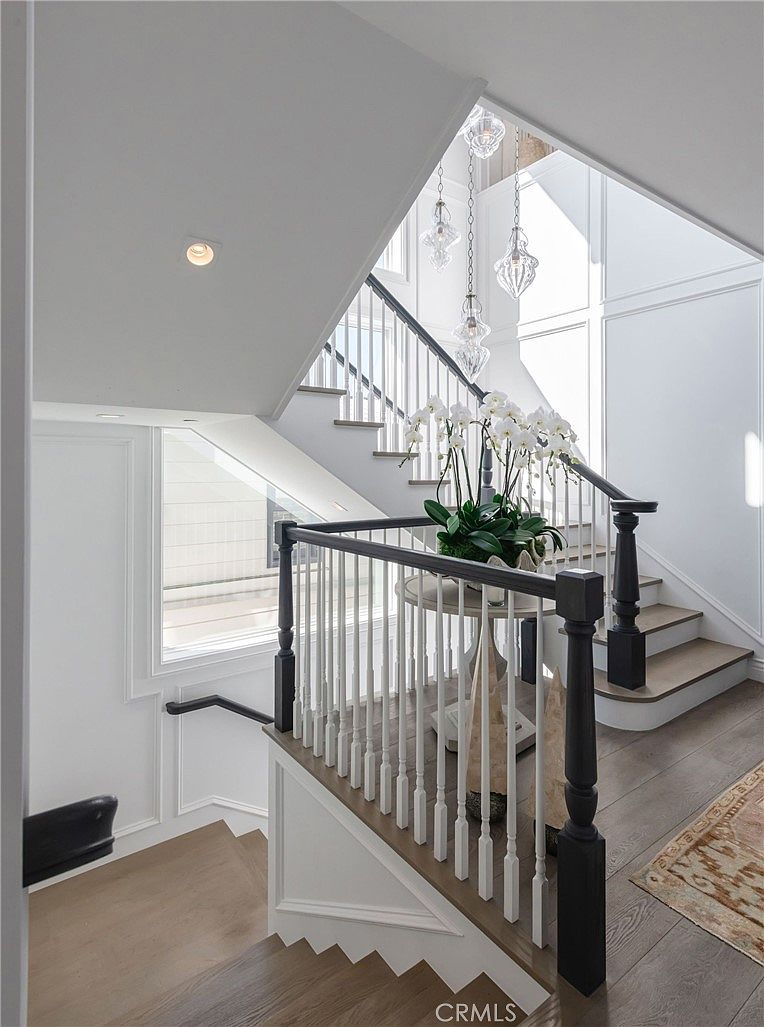 This elegant staircase features a classic design with white balusters, dark wood handrails, and matching dark wood treads. A central landing showcases a round table topped with a large, vibrant orchid arrangement, illuminated by a cluster of decorative glass pendant lights hanging from above. The space is bright and airy, characterized by crisp white walls with wainscoting and large windows that allow natural light to flood the stairwell.