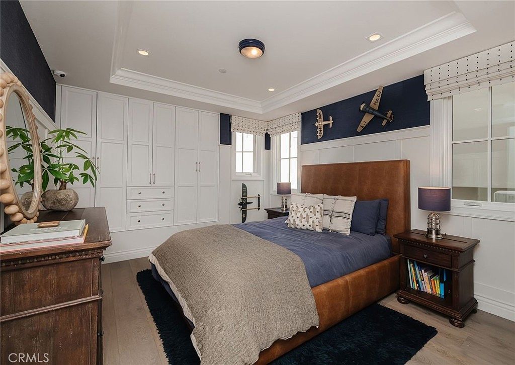 This well-appointed bedroom features a rich brown leather headboard paired with navy blue bedding, creating a sophisticated, aviation-themed aesthetic. The room includes built-in white cabinetry for ample storage, light wood flooring, and decorative model airplanes mounted on the navy-accented wall. The space is illuminated by a central ceiling fixture and bedside lamps, offering a cozy and organized atmosphere.
