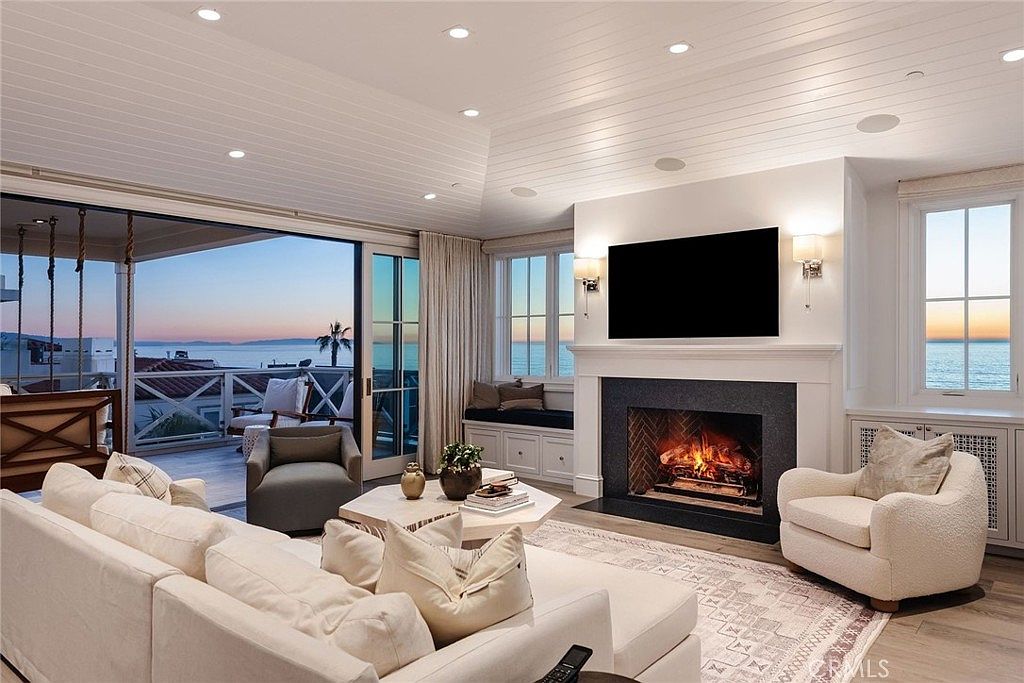 This elegant living room features a vaulted, shiplap-paneled ceiling and a large fireplace with a dark stone surround as its focal point. A plush, light-colored sectional sofa and a textured armchair provide comfortable seating, while a large sliding glass door opens to a balcony with expansive ocean views. The space is bright and airy, blending coastal charm with sophisticated interior design.