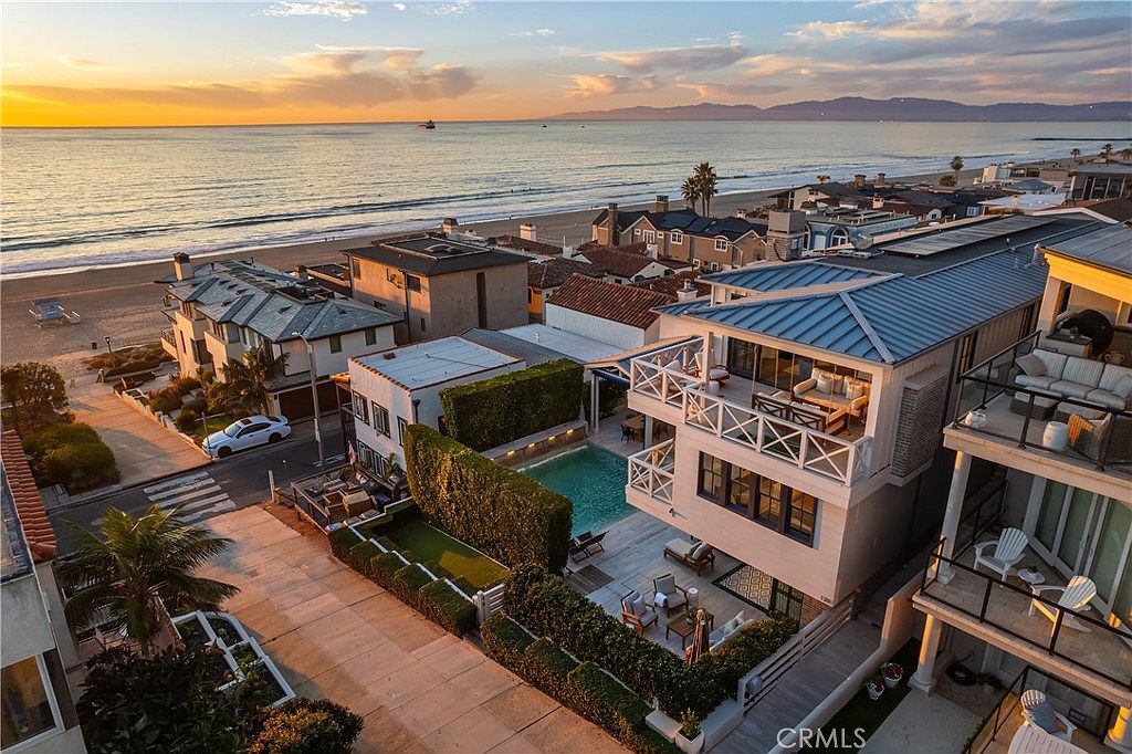 This high-angle aerial view captures a luxurious beachfront property at sunset, showcasing its prime location directly adjacent to the ocean. The home features a multi-level design with a private swimming pool, a spacious ground-level patio, and an expansive upper-level balcony with outdoor seating. The surrounding neighborhood is densely packed with coastal residences, all set against the backdrop of a vast, calm sea and distant mountains.