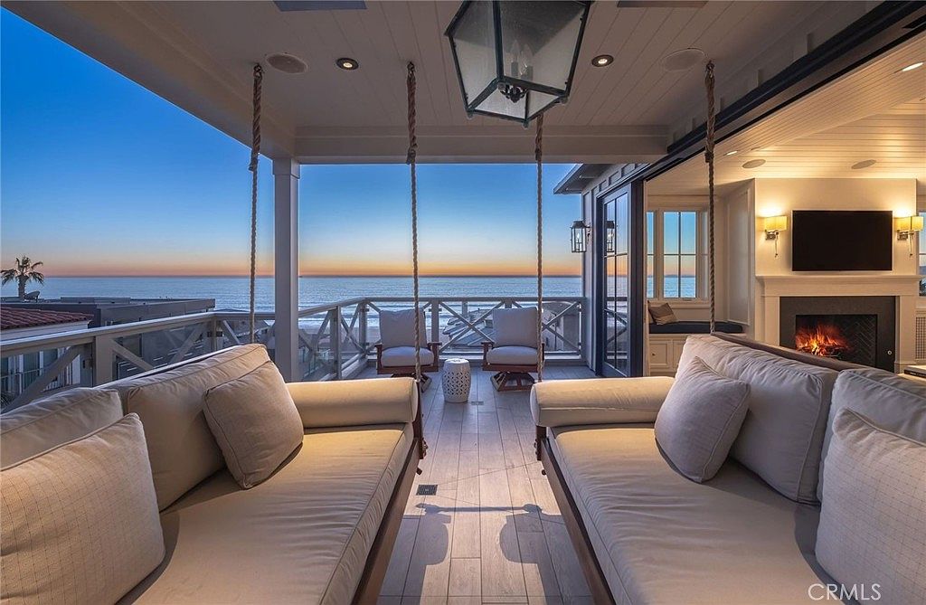 This luxurious covered balcony offers a seamless indoor-outdoor living experience with a stunning sunset view over the ocean. The space features two large, plush hanging daybeds facing each other, a central seating area with two chairs, and a cozy fireplace integrated into the adjacent indoor living space. The open-air design, combined with recessed lighting and high-end finishes, creates a sophisticated and tranquil atmosphere perfect for relaxation.