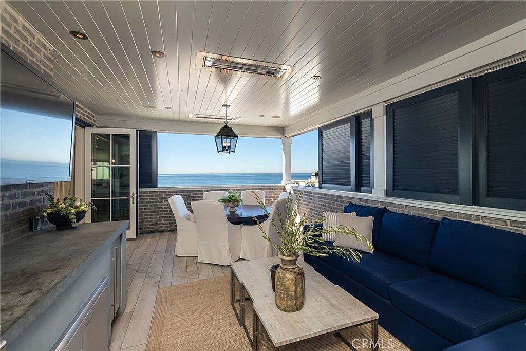 This covered outdoor living space offers a sophisticated, open-air lounge and dining area with a direct, unobstructed view of the ocean. The space features a navy blue sofa, a light wood coffee table, and a dining set, all set against a backdrop of brick walls and a paneled ceiling with integrated lighting. The open design seamlessly connects the indoor-outdoor transition, creating a serene and luxurious coastal retreat.