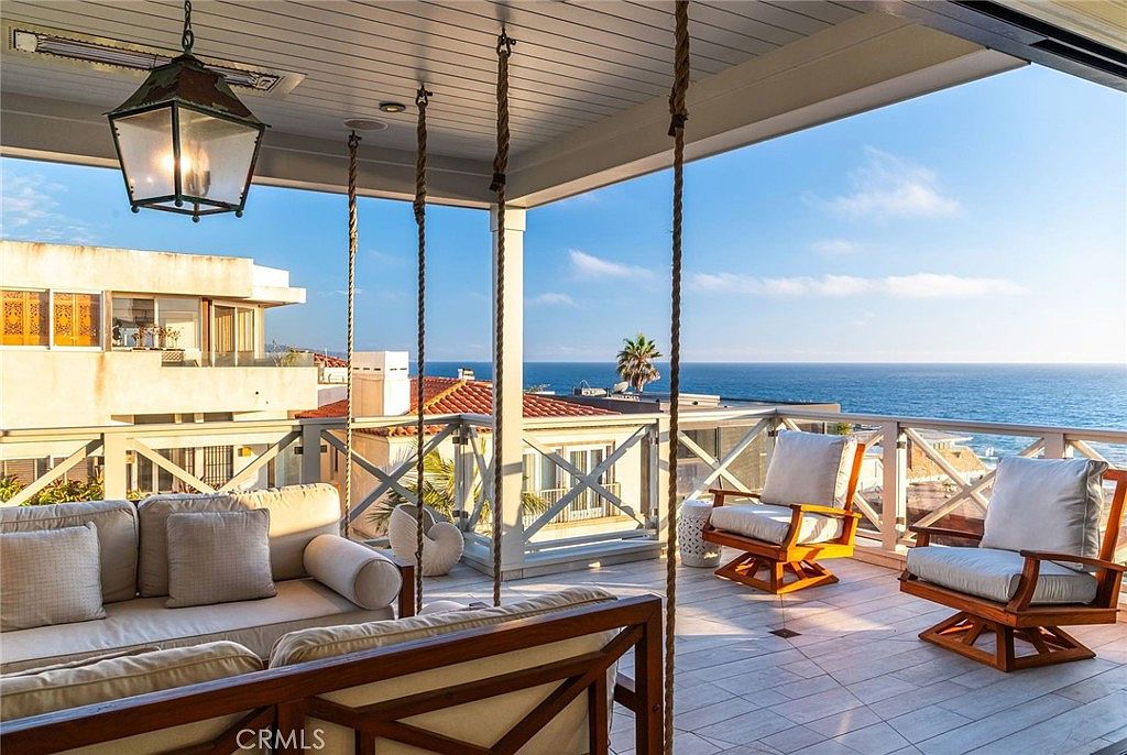This expansive, covered balcony offers a luxurious outdoor living space with a panoramic view of the ocean. The area is furnished with a comfortable sofa and two swivel chairs, all arranged to take advantage of the coastal scenery. A large, decorative lantern hangs from the ceiling, and the space is framed by a white railing, creating a serene and sophisticated atmosphere for relaxation.