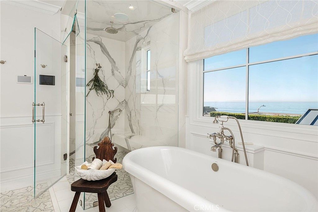 This luxurious primary bathroom features a freestanding soaking tub positioned beneath a large window that offers a serene view of the ocean. To the left, a glass-enclosed walk-in shower is finished with light-toned stone walls featuring prominent veining and a decorative patterned tile floor. The space is bright and airy, accented by a rustic wooden stool and elegant polished metal fixtures, creating a sophisticated coastal retreat.