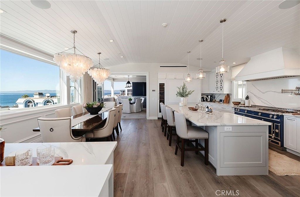 This bright, open-concept kitchen and dining area features a large central island with seating and a long dining table positioned beside expansive windows overlooking the ocean. The space is characterized by light wood flooring, white cabinetry, and elegant pendant lighting, creating a sophisticated and airy coastal aesthetic. The perspective captures the seamless flow between the culinary workspace and the dining area, emphasizing the panoramic water views.