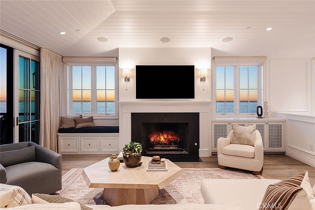 This elegant living room features a central fireplace with a dark surround and a mounted television, flanked by built-in cabinetry and window seats. Large windows on either side of the fireplace offer expansive ocean views, while a light-toned, geometric-patterned area rug anchors the seating area. The room is furnished with a light-colored sofa, a grey armchair, and a unique hexagonal coffee table, creating a sophisticated and serene coastal atmosphere.