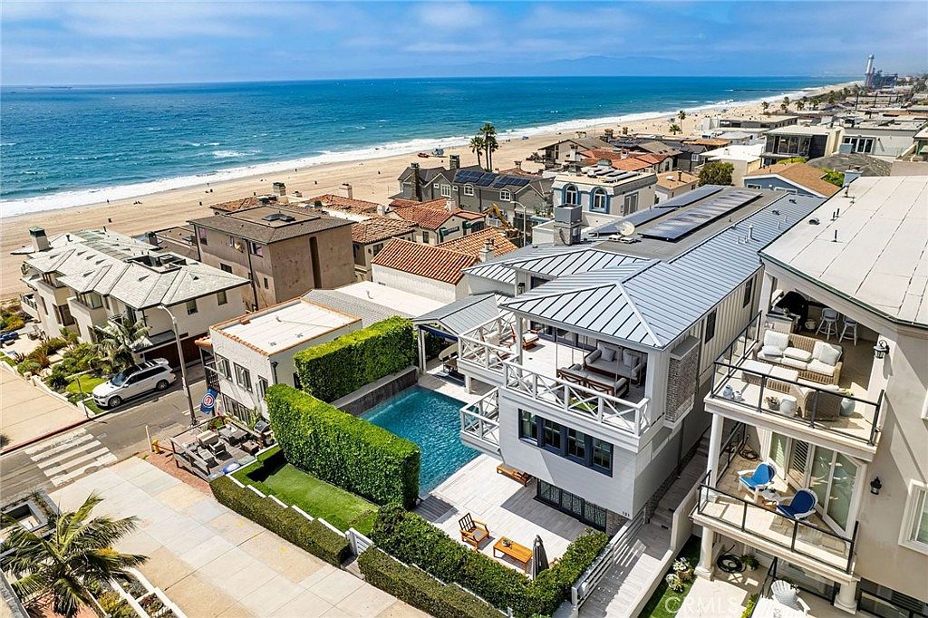 This high-angle aerial view captures a coastal property featuring a private swimming pool and multiple levels of outdoor living space, including a spacious balcony with lounge seating. The home is situated just steps from the sandy beach and ocean, showcasing a modern architectural style with a metal roof and clean lines. The surrounding neighborhood is densely packed with similar beach-front residences, emphasizing the prime location and proximity to the water.