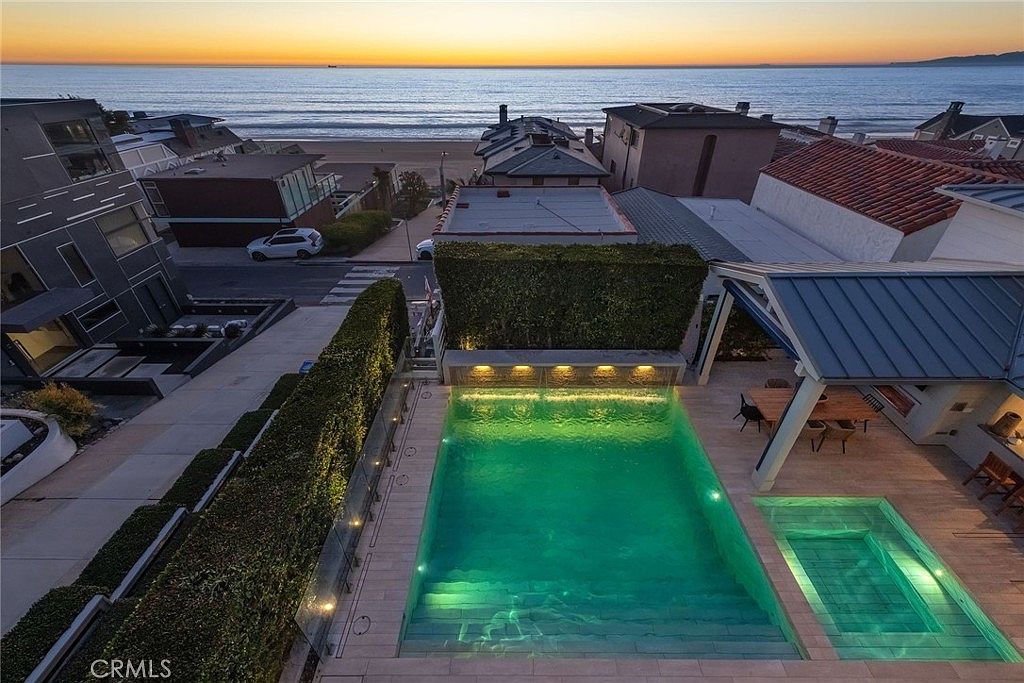 This high-angle aerial view captures a luxurious outdoor living space featuring a glowing, illuminated swimming pool and an adjacent spa. The pool area is bordered by a tall, manicured hedge for privacy and includes a covered patio space with outdoor dining furniture. Beyond the property, the scene overlooks a coastal street and the expansive ocean at sunset, creating a serene and upscale atmosphere.