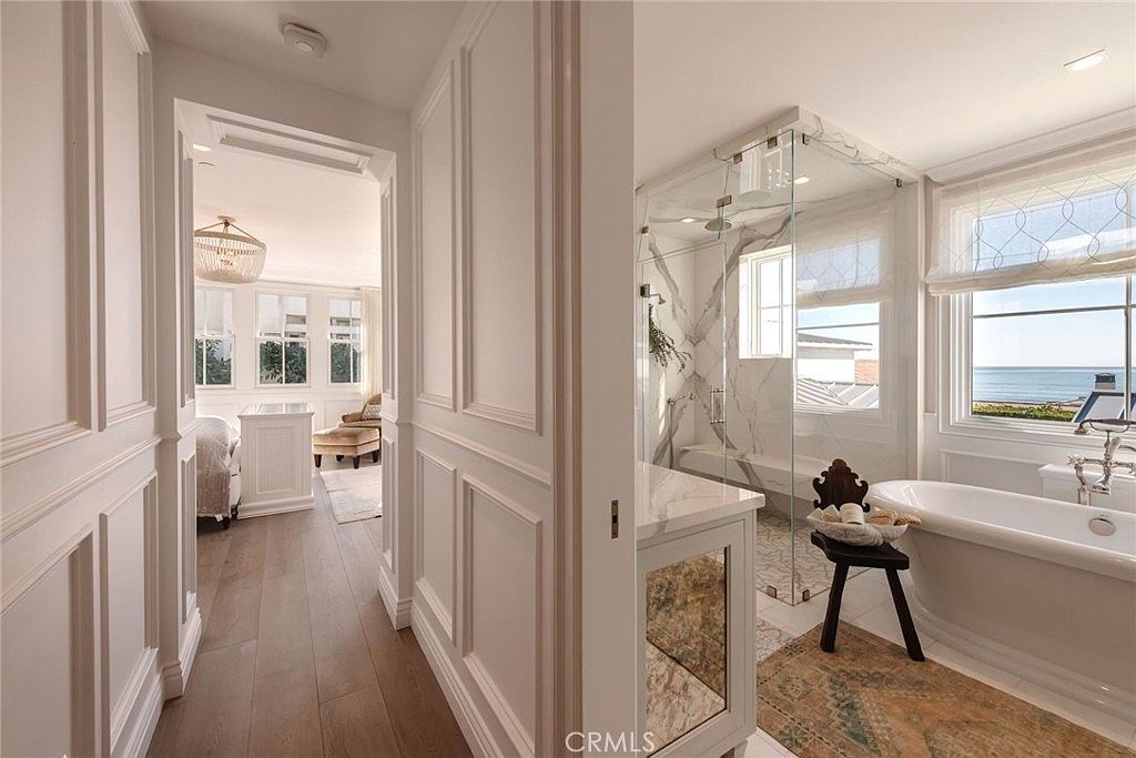 This view captures a luxurious primary bathroom featuring a freestanding soaking tub positioned next to a large window with ocean views. A glass-enclosed walk-in shower with light-toned stone walls sits adjacent to a vanity with a mirrored cabinet, while a hallway with paneled walls leads into an adjacent bedroom. The space is bright and airy, characterized by elegant millwork and a sophisticated, coastal-inspired aesthetic.