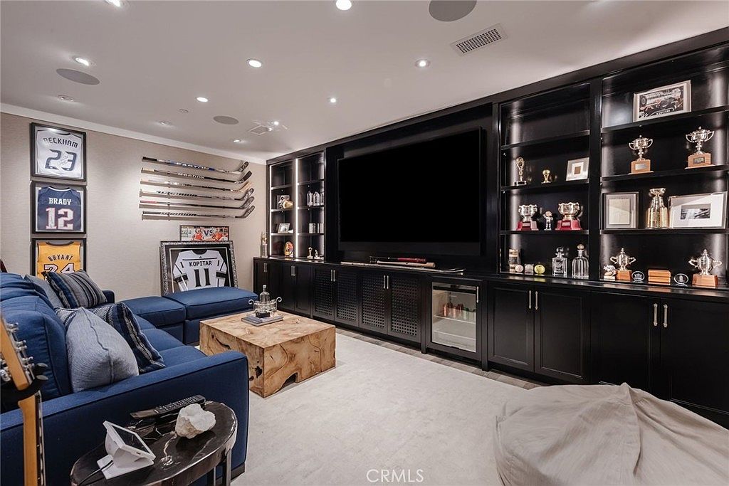 This sophisticated media room features a large wall-mounted television integrated into a custom black built-in shelving unit that displays various sports memorabilia and trophies. A plush blue sectional sofa provides ample seating, complemented by a unique natural wood coffee table and a soft, light-colored area rug. The room is decorated with framed sports jerseys on the wall, creating a dedicated space for entertainment and relaxation.