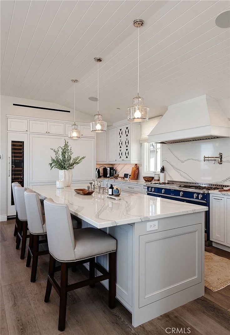 This bright, modern kitchen features a large central island with stone countertops and seating for three, illuminated by three glass pendant lights. The space is anchored by a striking blue range with a custom hood and a light-toned stone backsplash, complemented by white cabinetry and light wood flooring. The perspective captures the functional elegance of the workspace, highlighting the clean lines and high-end finishes that define this sophisticated culinary area.