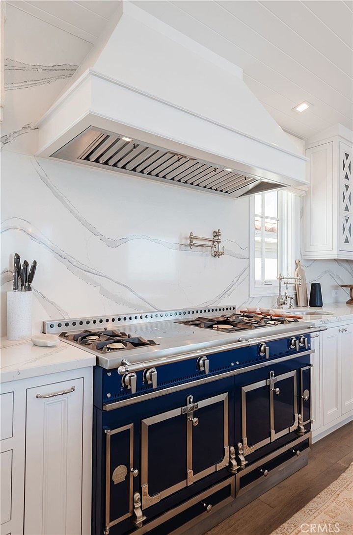 This high-end kitchen features a striking navy blue professional-style range with metallic accents, positioned beneath a large, custom-built white range hood. The backsplash is composed of large-format stone slabs with dramatic grey veining, complemented by a wall-mounted pot filler faucet. The perspective captures the cooking station with white cabinetry and light-toned stone countertops, creating a sophisticated and luxurious culinary space.