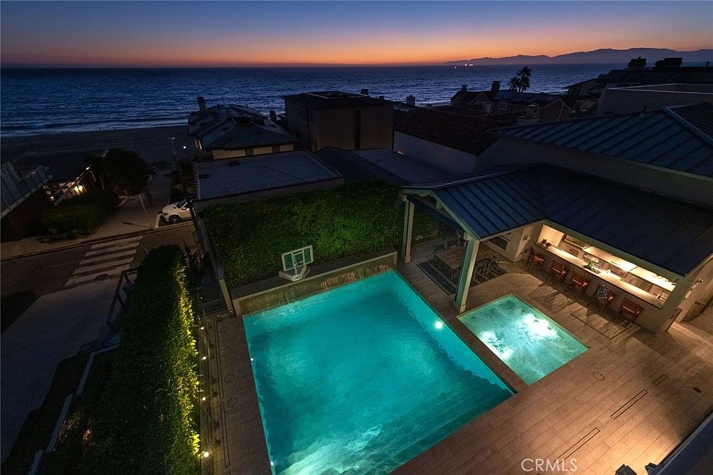 This high-angle aerial view captures a luxurious coastal backyard at twilight, featuring a glowing swimming pool and an adjacent spa. A covered outdoor lounge area with a bar and seating provides a perfect vantage point for the ocean sunset in the background. The scene is framed by lush greenery and modern hardscaping, creating an inviting and sophisticated atmosphere for evening relaxation.