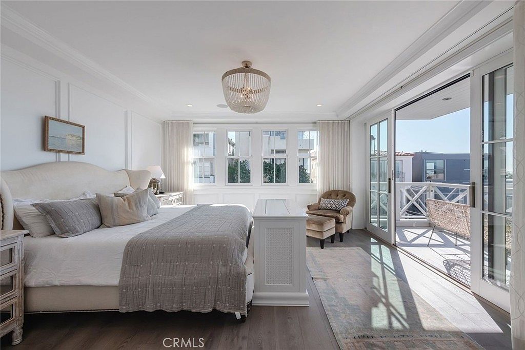 This bright and airy primary bedroom features a large upholstered bed with neutral bedding and a textured gray throw. The room is illuminated by a decorative beaded chandelier and natural light streaming through a row of windows and a set of glass doors that open onto a private balcony. A comfortable armchair sits in the corner, and the space is finished with dark wood flooring and a patterned area rug, creating a serene and sophisticated atmosphere.
