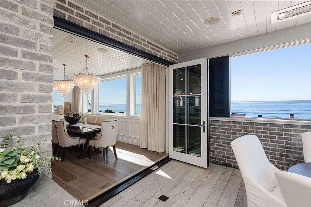This bright, open-concept space seamlessly blends an indoor dining area with an adjacent covered patio, both offering expansive ocean views. The room features light-toned wood flooring, a white-painted wood-paneled ceiling, and a distinctive gray brick wall that adds texture to the coastal aesthetic. Large glass doors open fully to connect the interior dining room with the outdoor seating area, creating an airy, indoor-outdoor living experience.