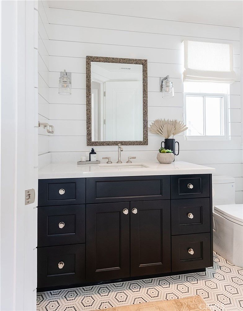 This bright bathroom features a dark vanity with a light-colored countertop and a framed mirror, set against white horizontal wall paneling. The space is accented by two wall-mounted light fixtures and a patterned tile floor, creating a clean, modern farmhouse aesthetic. A window with a fabric shade allows natural light to enter, while a decorative plant adds a touch of greenery to the vanity surface.