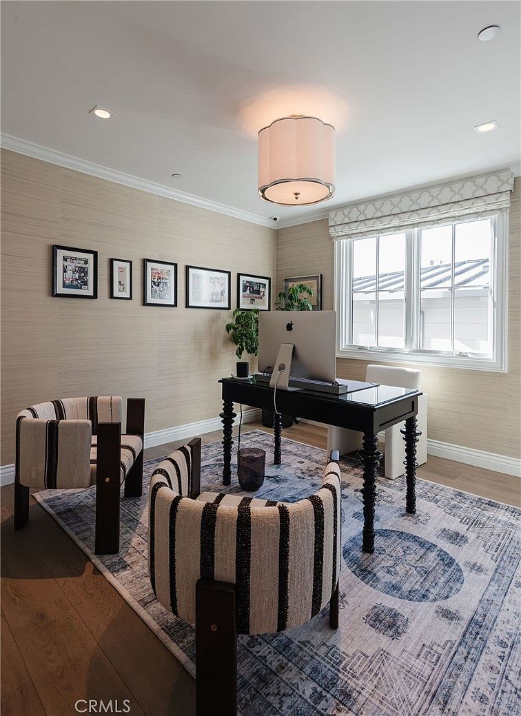 This sophisticated home office features a dark wood desk centered on a large, patterned area rug, flanked by two striped armchairs. The walls are adorned with textured wallpaper and a gallery wall of framed art, while a drum-style pendant light hangs above. A window with tailored roman shades provides natural light, creating a professional yet inviting workspace.