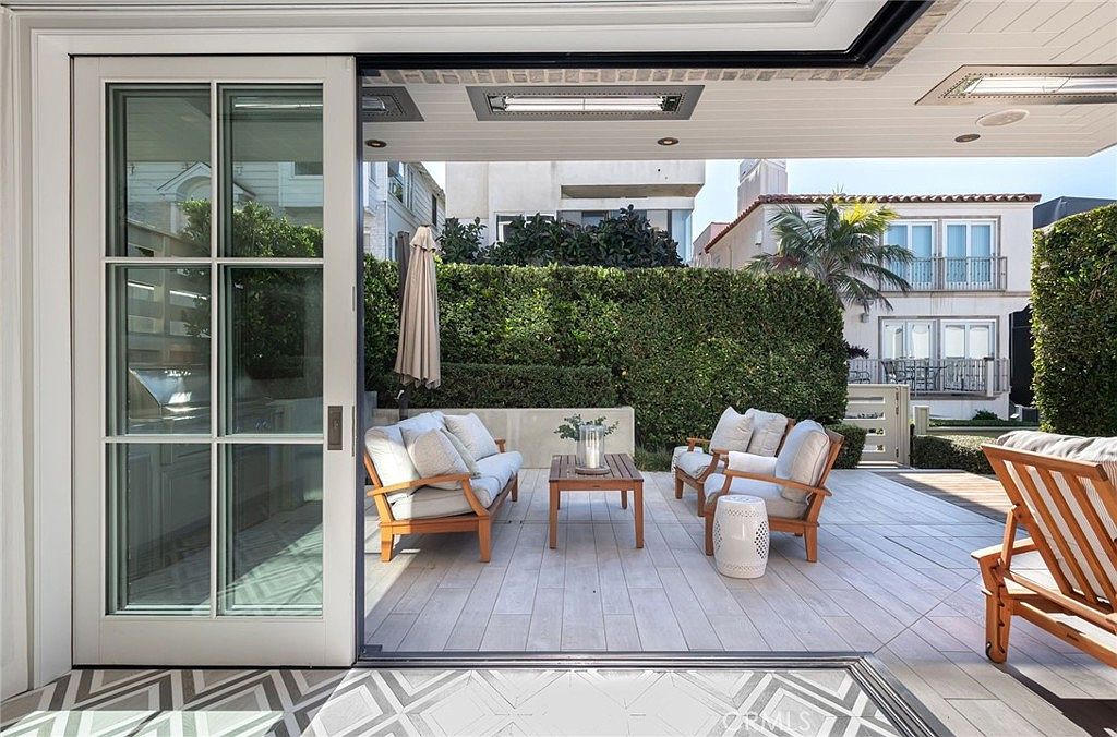 This image captures a seamless transition from an indoor space to a spacious outdoor patio, framed by large sliding glass doors. The patio is furnished with a comfortable seating arrangement, including a sofa and armchairs with light-colored cushions, centered around a small wooden coffee table. A tall, manicured hedge provides privacy, while the light-toned tiled flooring and overhead heaters create an inviting, sophisticated atmosphere for outdoor relaxation.