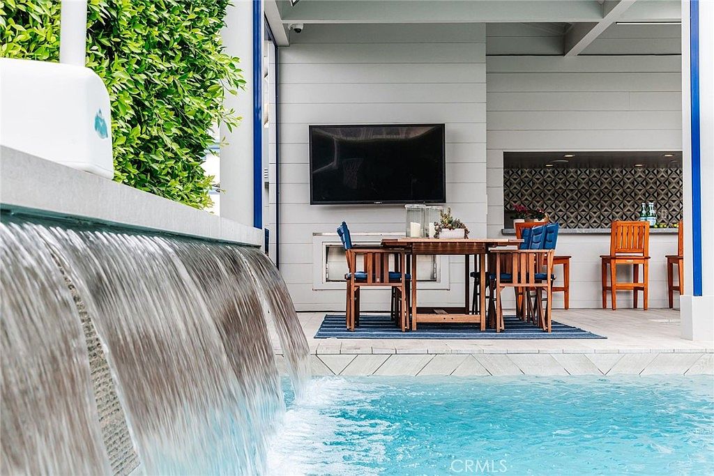 This outdoor living space features a modern swimming pool with a prominent water feature cascading over a stone wall. Adjacent to the pool is a covered patio area equipped with a wall-mounted television, a dining set, and a built-in outdoor kitchen area with patterned tile backsplash. The space is designed for seamless indoor-outdoor entertaining, offering a sophisticated and relaxing atmosphere.
