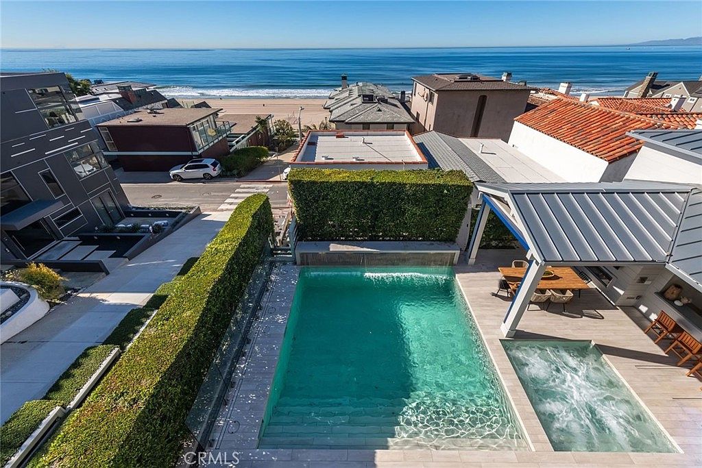 This high-angle aerial view captures a luxurious coastal backyard featuring a rectangular swimming pool with a cascading water feature and an adjacent square spa. The pool area is complemented by a covered patio space with outdoor seating, all set against a backdrop of a sandy beach and the expansive ocean. The scene conveys a high-end, serene lifestyle with clean lines and a seamless transition between the private outdoor living space and the natural coastal environment.