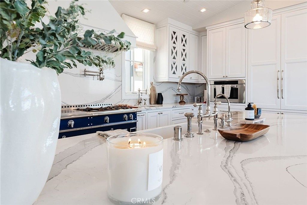 This bright, high-end kitchen features a large central island with a stone countertop and a polished metal faucet, foregrounded by a lit candle. A striking blue range with a matching hood serves as a focal point against a backdrop of white cabinetry and patterned stone backsplash. The space is illuminated by recessed lighting and a glass pendant, creating a clean, sophisticated, and airy atmosphere.