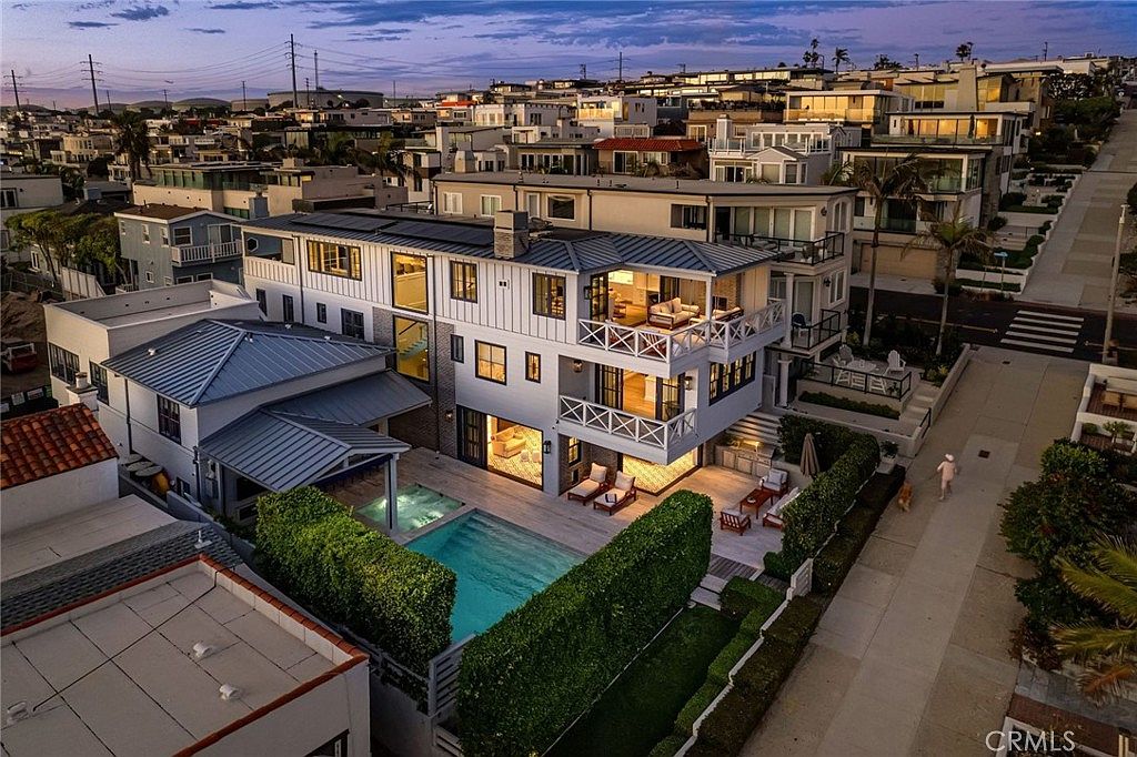 This high-angle aerial shot captures a modern, multi-level coastal residence at dusk, showcasing its contemporary architectural design and integrated outdoor living spaces. The property features a private swimming pool and spa, multiple levels of balconies with white railings, and large glass sliding doors that open to the patio. The surrounding neighborhood is densely packed with similar upscale homes, and the scene is illuminated by warm interior lighting that contrasts with the cool twilight sky.