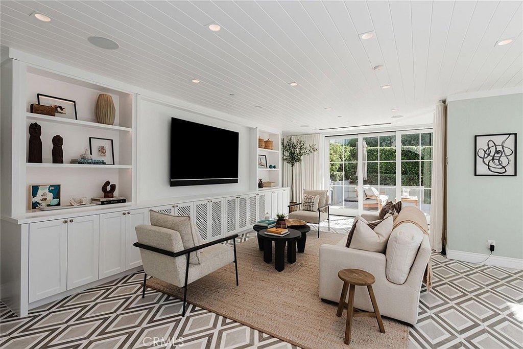 This bright and airy living room features custom built-in cabinetry surrounding a large wall-mounted television, complemented by a shiplap ceiling with recessed lighting. The space is furnished with a neutral-toned sofa, two armchairs, and a pair of round coffee tables, all anchored by a textured area rug over a geometric-patterned floor. Large glass sliding doors provide a seamless transition to the outdoor patio, filling the room with natural light and creating an inviting, sophisticated atmosphere.