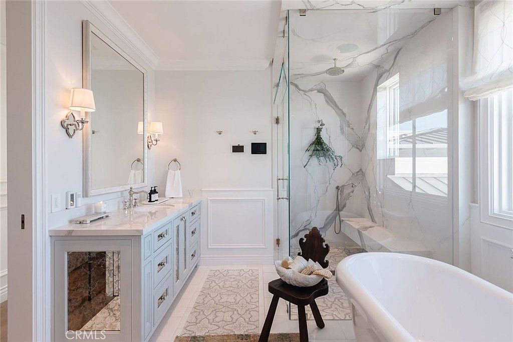 This elegant primary bathroom features a double vanity with a large mirror, a freestanding soaking tub, and a spacious walk-in shower enclosed by glass panels. The space is finished with light-toned cabinetry, stone surfaces, and a patterned tile floor, creating a bright and luxurious atmosphere. The perspective captures the entire room, highlighting the seamless transition between the vanity area and the wet room.