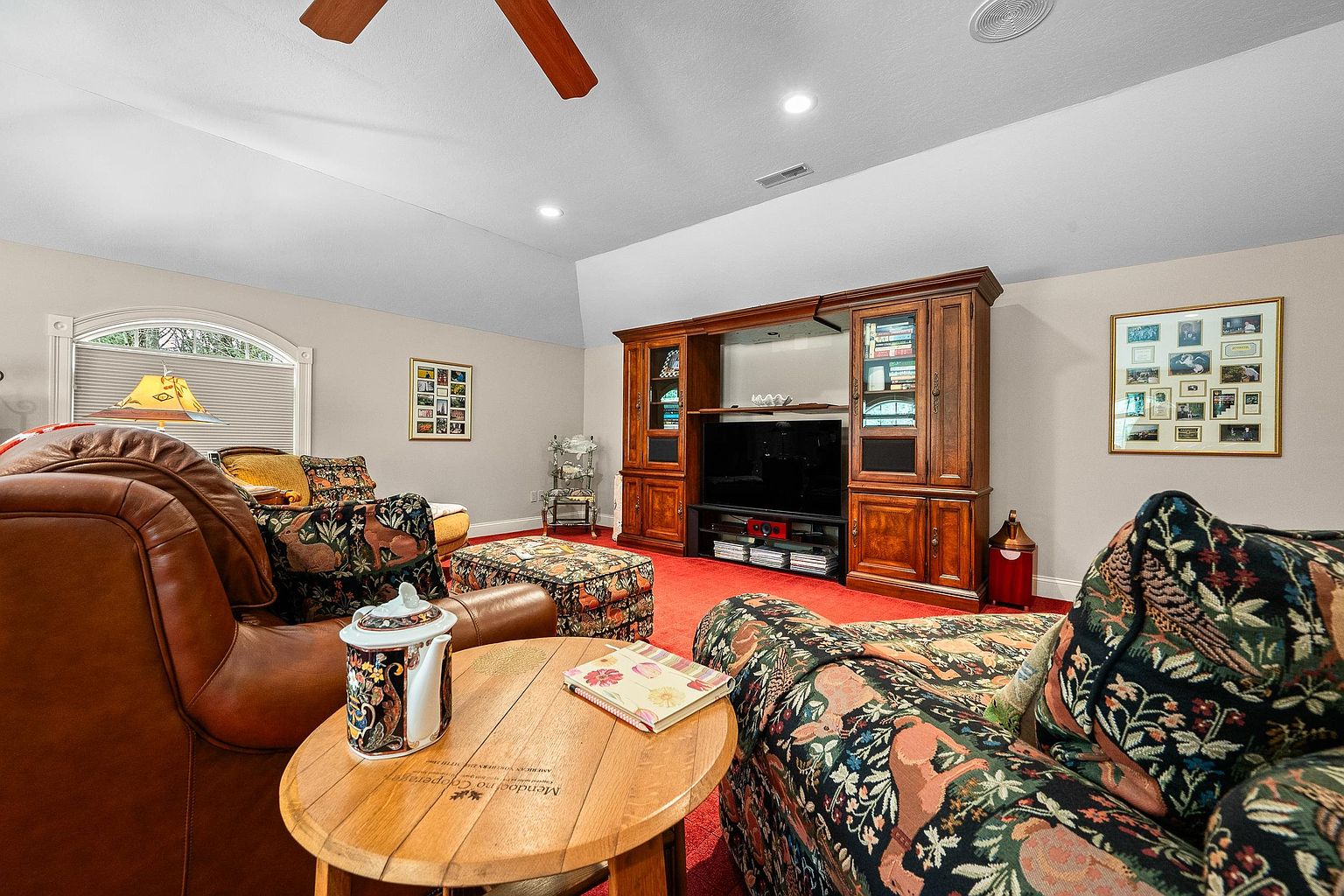 This cozy living room features a traditional aesthetic with a large, ornate wooden entertainment center as the focal point. The space is furnished with plush, patterned upholstery and a leather sofa, all set against a vibrant red carpet. A round wooden side table in the foreground holds a decorative teapot and a book, creating a warm and inviting atmosphere.