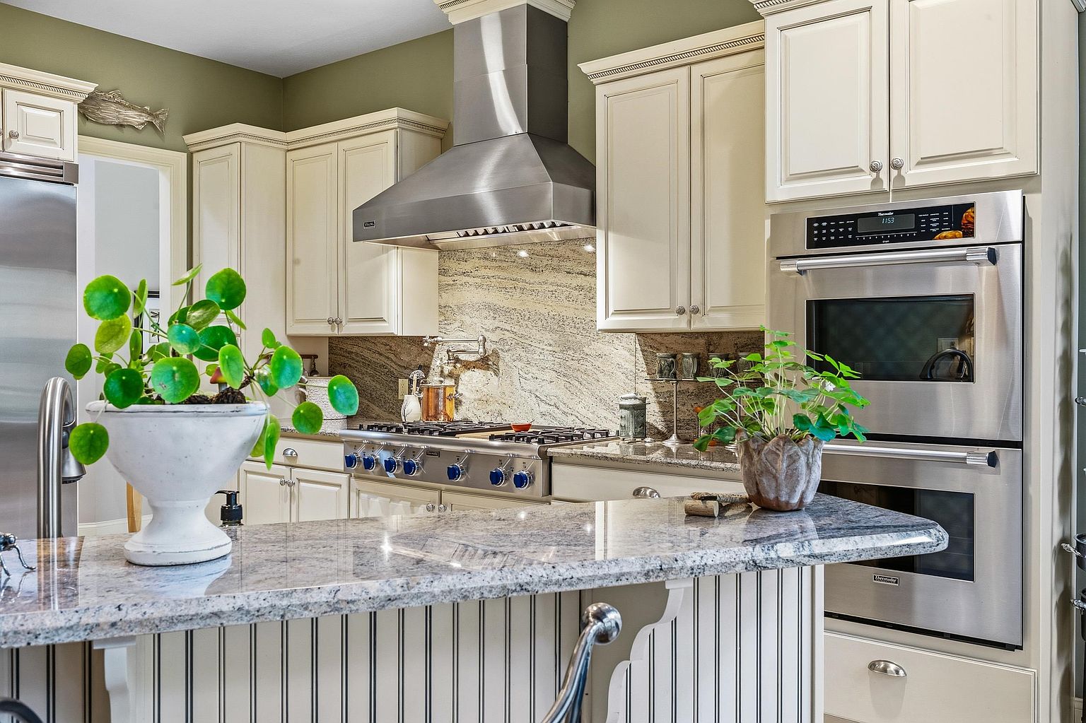 This elegant kitchen features cream-colored cabinetry, a professional-grade stainless steel range with a matching hood, and a striking granite countertop island. The space is accented by a decorative pinstriped base on the island, a natural stone backsplash, and lush potted plants that add a touch of warmth to the sophisticated design. The perspective is a eye-level shot capturing the functional layout and high-end finishes, creating an inviting and upscale atmosphere.