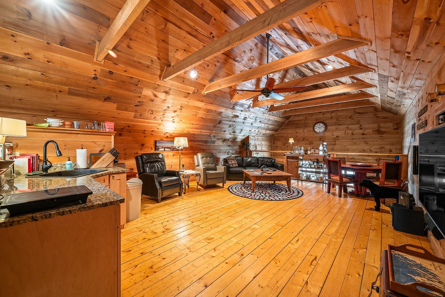 This spacious, open-concept living area features warm wood-paneled walls and vaulted ceilings with exposed beams, creating a cozy, rustic cabin atmosphere. The room includes a functional kitchen area with granite countertops, a comfortable seating arrangement with leather furniture, and a dedicated dining space with a bar cart. The perspective is a wide-angle shot from the kitchen, showcasing the seamless flow between the cooking, lounging, and dining zones.