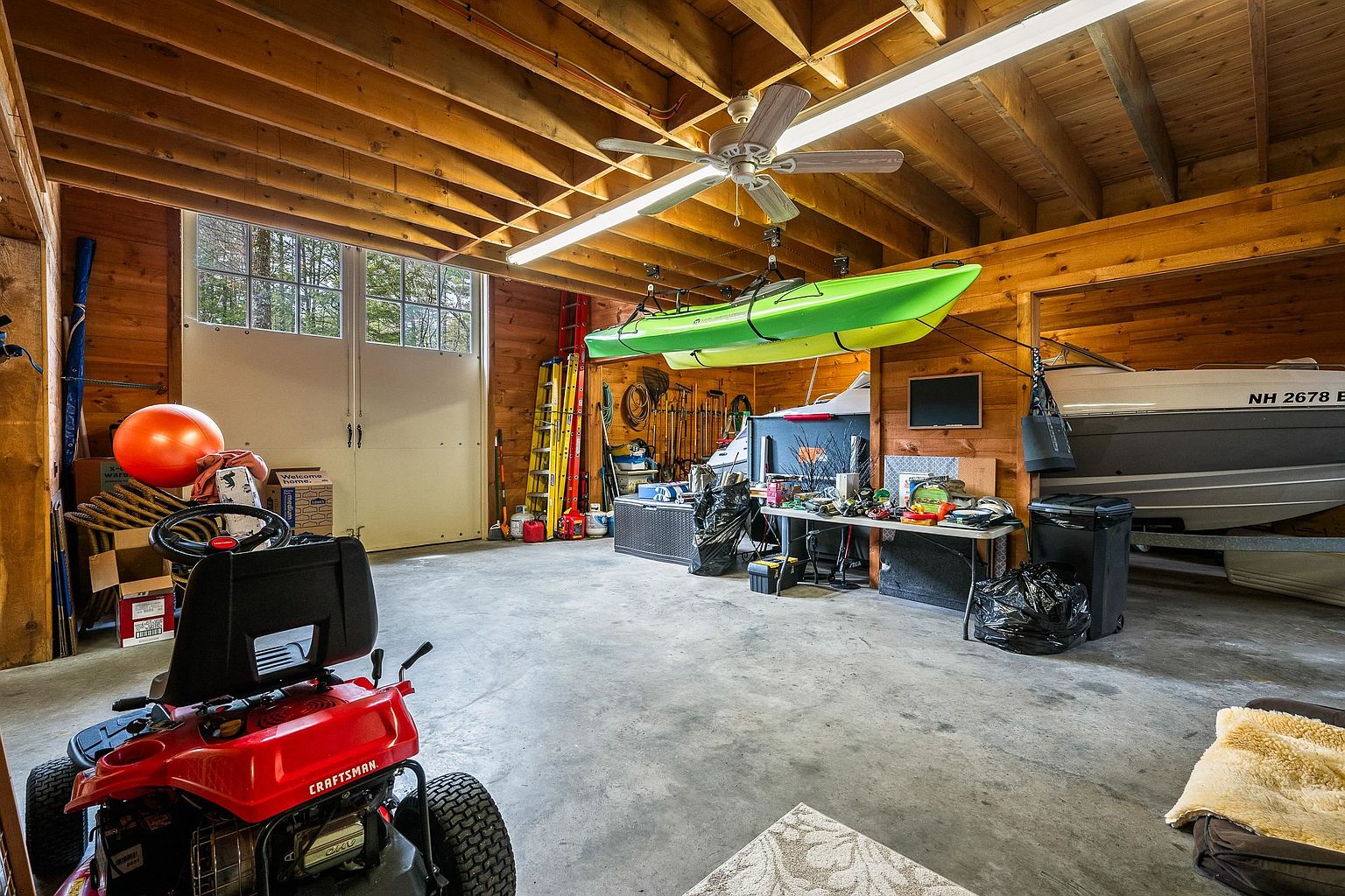 This spacious, rustic-style garage features exposed wooden ceiling beams, concrete flooring, and large double doors that allow for natural light. The area is well-equipped for storage and hobbies, housing a red Craftsman riding lawnmower, a suspended bright green kayak, and a boat parked in a dedicated bay. The overall impression is that of a functional, versatile workshop space perfect for outdoor enthusiasts.