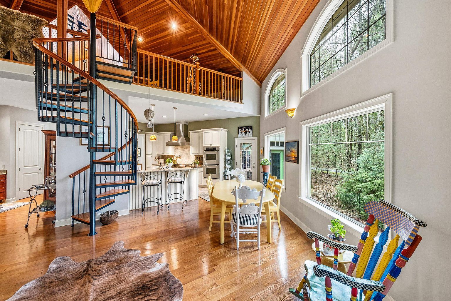 This open-concept living and dining area features a striking spiral staircase and a soaring vaulted ceiling with warm wood paneling. Large windows flood the space with natural light, highlighting the hardwood floors and the colorful, eclectic rocking chair in the foreground. The kitchen is visible in the background, creating a cohesive and inviting atmosphere that blends rustic charm with modern living.