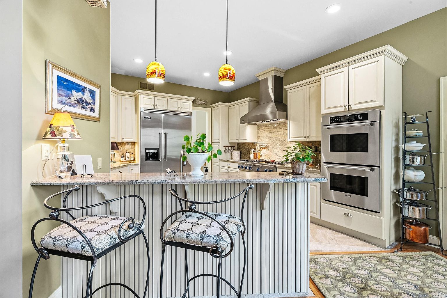 This elegant kitchen features a breakfast bar with patterned stools, granite countertops, and cream-colored cabinetry. The space is well-appointed with stainless steel appliances, including a double wall oven and a large refrigerator, complemented by warm pendant lighting. The overall aesthetic is traditional and inviting, with a decorative rug and a tiered pot rack adding character to the functional layout.