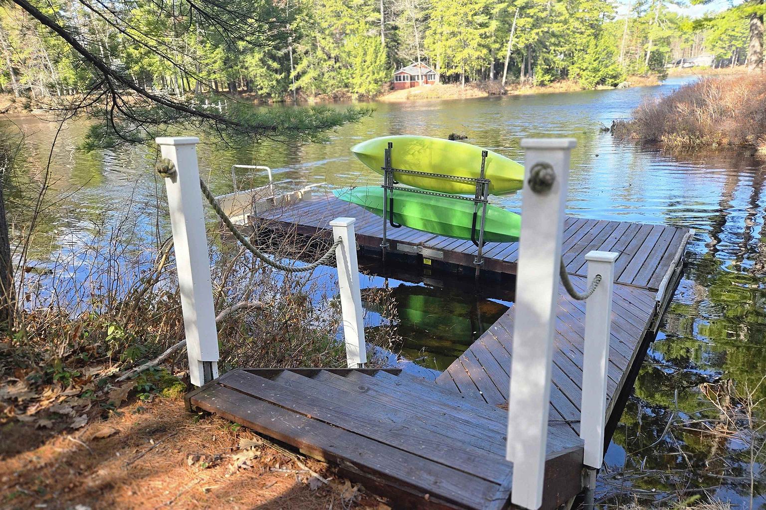 This image captures a serene waterfront scene featuring a wooden dock extending into a calm lake, complete with a rack holding two kayaks. The perspective is from the shore, looking down a short set of wooden stairs that lead directly onto the dock, framed by white posts and rope railings. In the background, a small cabin is nestled among lush green trees, creating a peaceful and inviting lakeside retreat.
