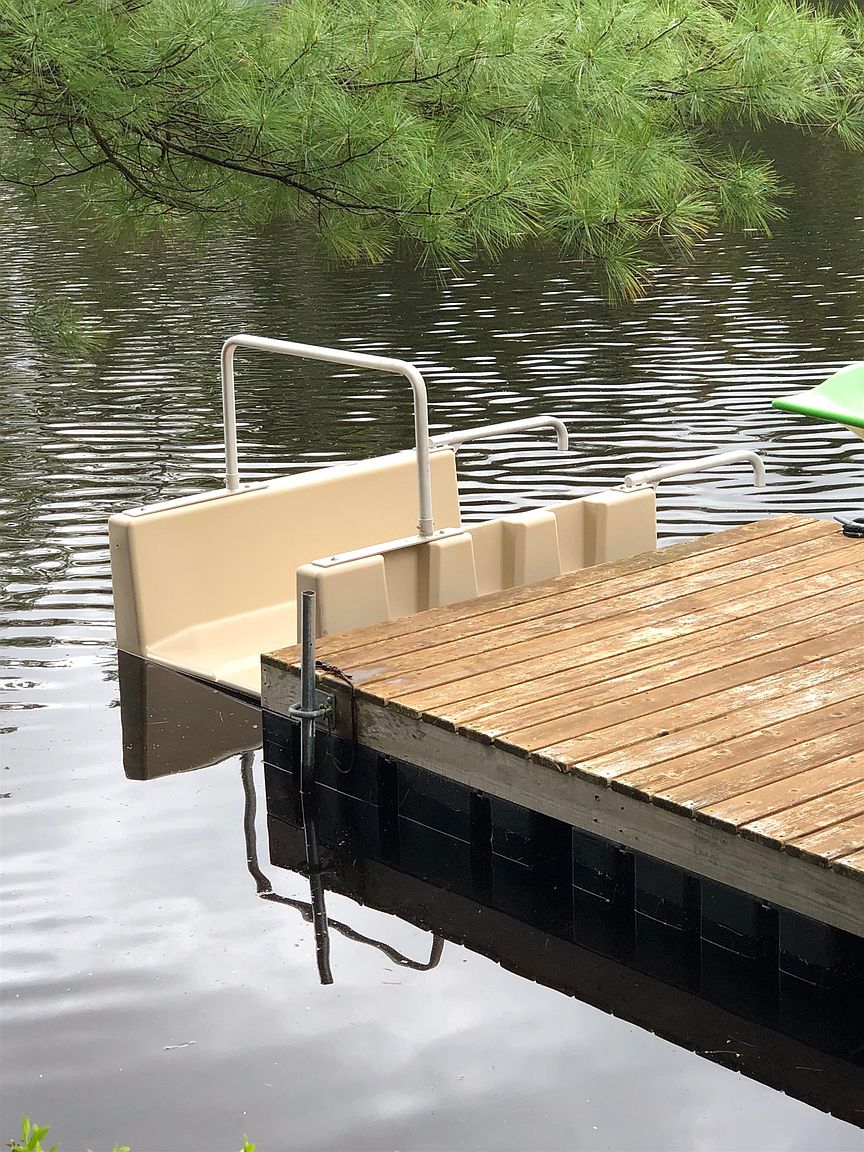 This image captures a close-up, eye-level perspective of a wooden dock extending over a calm lake, featuring a specialized beige plastic swim ladder or boarding platform with metal handrails. The scene is framed by the overhanging green needles of a pine tree, creating a peaceful and rustic waterfront atmosphere. The focus on the functional dock equipment highlights the property's direct access to the water, ideal for recreational activities.