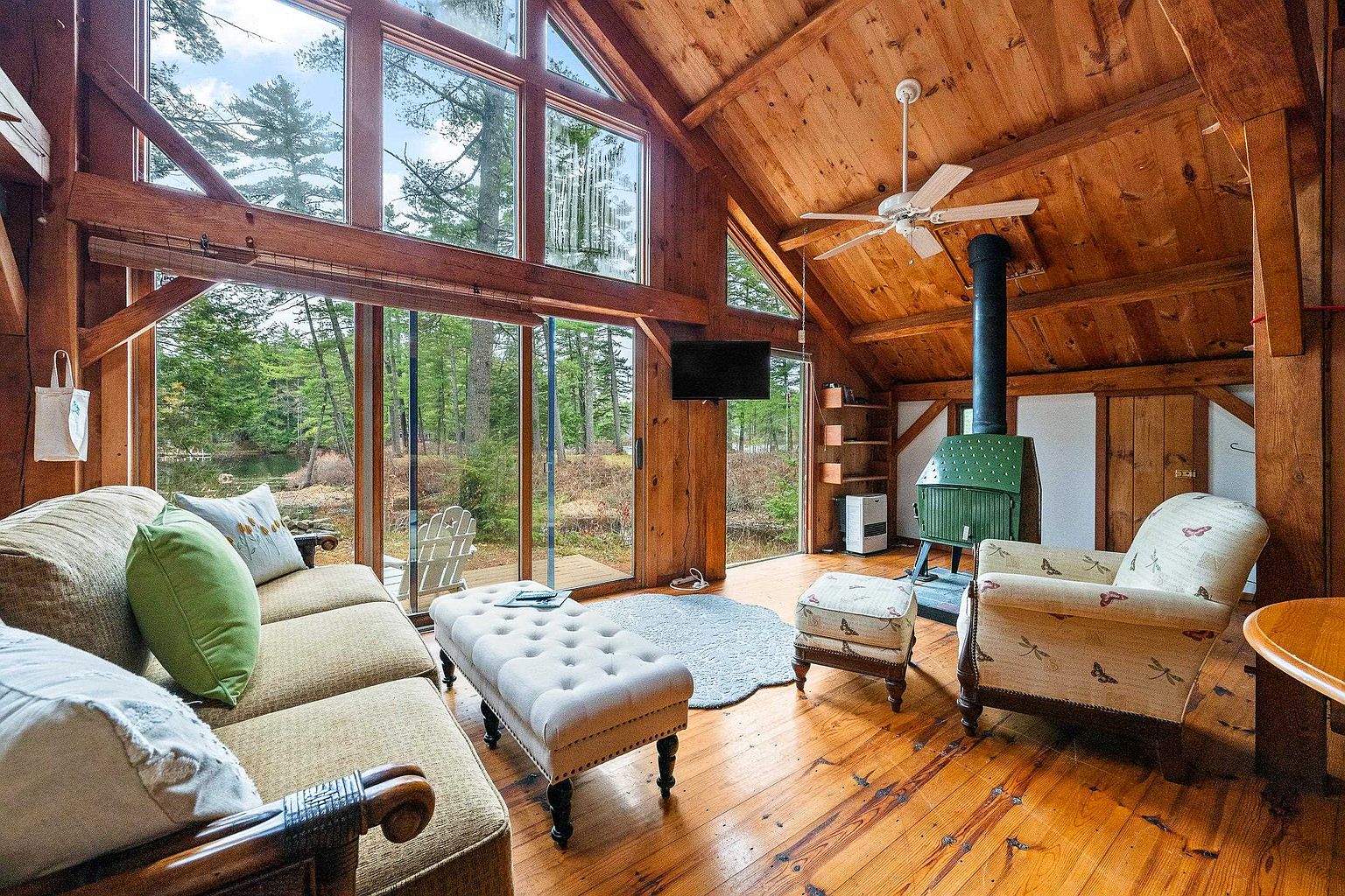 This cozy, rustic living room features warm wood-paneled walls and ceilings, complemented by a large floor-to-ceiling window wall that offers a serene view of the surrounding forest. The space is furnished with a comfortable sofa, a tufted ottoman, and a patterned armchair, all centered around a distinctive green wood-burning stove. The warm, natural lighting and timber construction create an inviting, cabin-like atmosphere perfect for relaxation.