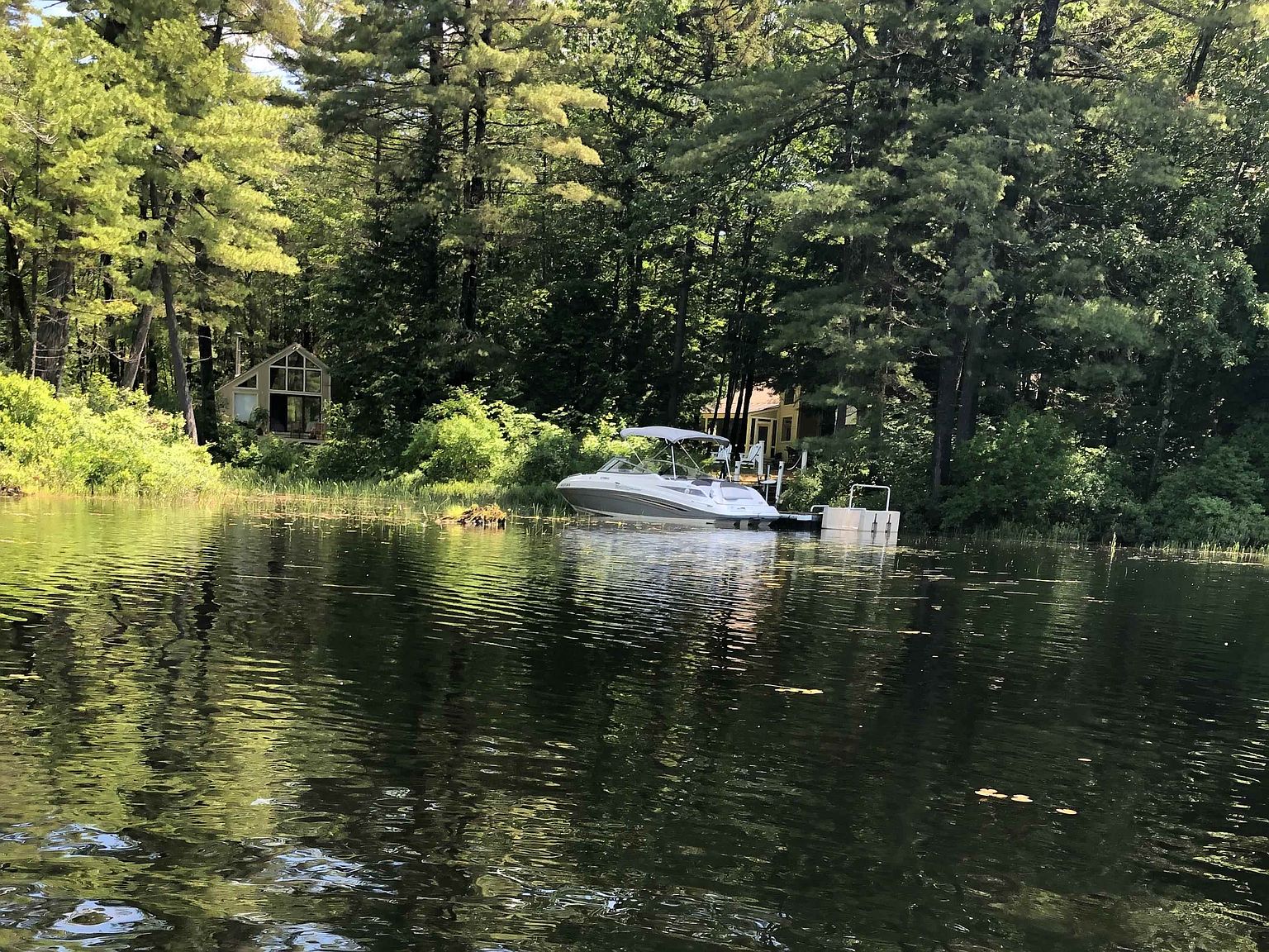This serene waterfront scene captures a tranquil lake setting featuring a modern cabin nestled among lush, mature pine trees. A sleek motorboat is docked at a private pier, emphasizing the recreational lifestyle associated with this property. The perspective is from the water looking toward the shoreline, highlighting the peaceful integration of the home into its natural, wooded environment.