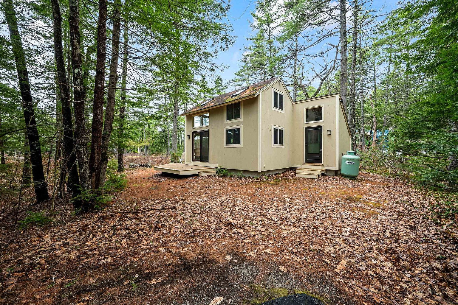 This image captures a charming, compact cabin nestled within a dense, wooded forest setting. The structure features light-colored siding, a small wooden deck, and a prominent propane tank near the side entrance, evoking a peaceful and secluded retreat atmosphere. The perspective is a wide-angle shot from the forest floor, emphasizing the home's integration into the natural, leaf-covered landscape.