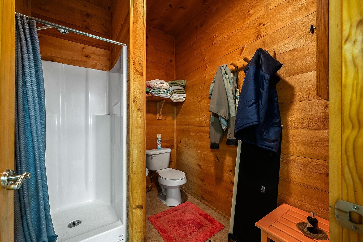 This rustic-style bathroom features warm wood-paneled walls and a compact layout that maximizes space. It includes a white shower stall, a standard toilet, and a small wooden bench, creating a cozy and functional atmosphere. The perspective is from the doorway, looking into the small, efficient washroom area.