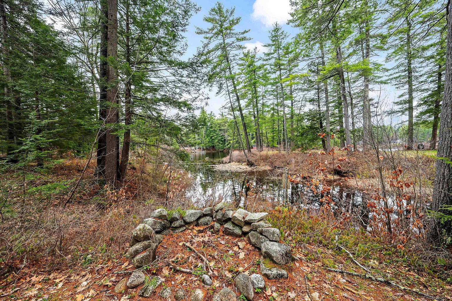 This scenic outdoor space features a rustic stone fire pit in the foreground, set against a backdrop of a tranquil, wooded pond. The natural landscape is filled with tall pine trees and dense brush, creating a peaceful and secluded atmosphere. The perspective is eye-level, capturing the serene connection between the property's grounds and the surrounding wilderness.